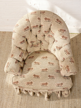 Load image into Gallery viewer, Image 6 of 18: Vintage upholstered barrel chair featuring beige textured fabric with repeating brown lion motifs, a deeply tufted rounded backrest, padded rolled arms, thick seat cushion, and matching ruffled skirt.
