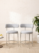 Load image into Gallery viewer, Image 1 of 18: Modern upholstered bar stool featuring a curved beige textured fabric backrest, cushioned seat, slender chrome-finished metal legs, and circular metal footrest. Sold as a pair.
