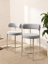 Load image into Gallery viewer, Image 2 of 18: Modern upholstered bar stool featuring a curved beige textured fabric backrest, cushioned seat, slender chrome-finished metal legs, and circular metal footrest. Sold as a pair.
