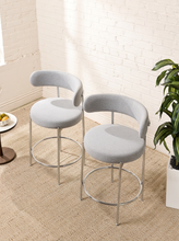 Load image into Gallery viewer, Image 3 of 18: Modern upholstered bar stool featuring a curved beige textured fabric backrest, cushioned seat, slender chrome-finished metal legs, and circular metal footrest. Sold as a pair.
