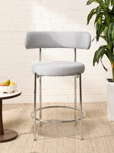 Load image into Gallery viewer, Image 4 of 18: Modern upholstered bar stool featuring a curved beige textured fabric backrest, cushioned seat, slender chrome-finished metal legs, and circular metal footrest. Sold as a pair.
