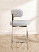 Load image into Gallery viewer, Image 7 of 18: Modern upholstered bar stool featuring a curved beige textured fabric backrest, cushioned seat, slender chrome-finished metal legs, and circular metal footrest. Sold as a pair.
