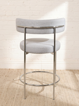 Load image into Gallery viewer, Image 8 of 18: Modern upholstered bar stool featuring a curved beige textured fabric backrest, cushioned seat, slender chrome-finished metal legs, and circular metal footrest. Sold as a pair.
