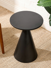 Load image into Gallery viewer, Image 6 of 14: A round black side table with a tapered cone base, shown on a textured beige rug in a room setting with a white brick wall and a green plant leaf visible.
