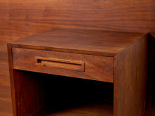 Load image into Gallery viewer, Image 11 of 22: A close-up of a mid-century modern wooden nightstand with a single drawer featuring a recessed pull handle, set against a matching wood-paneled background.
