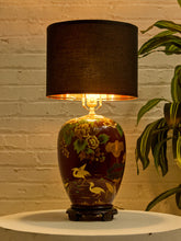 Load image into Gallery viewer, Image 2 of 19: Vintage 1950s table lamp featuring a rounded burgundy base with hand-painted yellow and cream flowers, green foliage, and decorative bird motifs, resting on a dark pedestal base and topped with a black cylindrical fabric shade.
