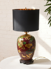 Load image into Gallery viewer, Image 3 of 19: Vintage 1950s table lamp featuring a rounded burgundy base with hand-painted yellow and cream flowers, green foliage, and decorative bird motifs, resting on a dark pedestal base and topped with a black cylindrical fabric shade.
