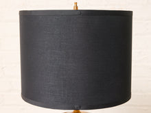 Load image into Gallery viewer, Image 10 of 19: A black fabric drum lampshade with a visible woven texture, shown against a light off-white brick wall background. Small brass finials are visible at the top and bottom of the lamp.
