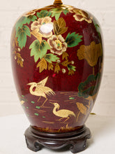 Load image into Gallery viewer, Image 14 of 19: A decorative ceramic vase with a glossy deep red glaze, decorated with hand-painted gold and green floral and bird motifs, displayed on a dark wooden stand against a white brick wall background.
