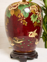 Load image into Gallery viewer, Image 15 of 19: A decorative ceramic vase with a glossy deep red body, hand-painted floral and bird motifs in gold, green, and beige, displayed on a dark wooden stand.
