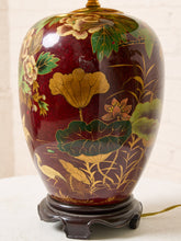 Load image into Gallery viewer, Image 16 of 19: A decorative ceramic table lamp with a glossy dark red body decorated with hand-painted gold and green lotus motifs, resting on a dark wooden base.
