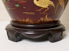 Load image into Gallery viewer, Image 18 of 19: Close-up of a decorative ceramic vase with a dark reddish-brown glaze, hand-painted gold crane and grass motifs, and a dark wooden stand.
