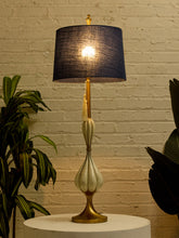 Load image into Gallery viewer, Image 2 of 19: A table lamp with a dark navy blue textured drum shade, a cream-colored sculpted ceramic body, and a gold-toned base, shown lit on a white pedestal against a white brick wall with plants.
