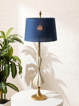 Load image into Gallery viewer, Image 3 of 19: A mid-century modern table lamp with a navy blue fabric drum shade, a cream-colored sculpted ceramic body, and a polished gold-tone base, displayed on a white pedestal against a white brick wall next to a green plant.

