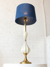 Load image into Gallery viewer, Image 5 of 19: A mid-century modern table lamp with a textured blue drum shade, a glossy white sculptural base, and a brass-toned metal stem and base, set against a white brick wall.
