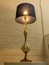 Load image into Gallery viewer, Image 6 of 19: A vintage-style table lamp with a dark navy blue fabric drum shade, a gold-toned metal neck, and a sculptural fluted base in cream and gold, illuminated against a white brick wall background.

