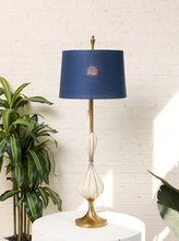 Load image into Gallery viewer, Image 7 of 19: A table lamp with a navy blue drum shade, a gold-finished metal stem and base, and a white ribbed glass neck, displayed on a white round pedestal against a white brick wall with a green plant to the side.
