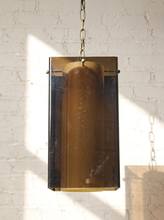 Load image into Gallery viewer, Image 3 of 24: A vintage rectangular pendant light with a translucent amber-brown glass shade and brass metal trim, hanging from a chain against a white brick wall with sunlight casting shadows.
