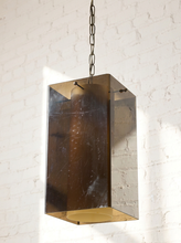 Load image into Gallery viewer, Image 5 of 24: A rectangular pendant light with a smoked amber glass shade and a brushed brass-toned metal frame, hanging from a dark chain against a white brick wall.
