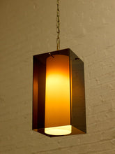 Load image into Gallery viewer, Image 6 of 24: A modern rectangular pendant light with a glowing warm yellow-orange frosted glass center and dark bronze metal sides, hanging from a chain against a white brick wall.
