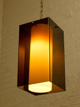 Load image into Gallery viewer, Image 8 of 24: A modern rectangular pendant light with a dark amber-brown translucent glass frame and a glowing warm yellow-orange cylindrical inner shade, hanging against a textured off-white brick wall.
