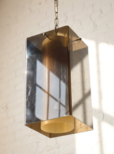 Load image into Gallery viewer, Image 7 of 24: A modern rectangular pendant light with a brushed silver metal frame and a warm brass cylindrical shade, hanging against a white brick wall with window shadow patterns.
