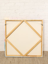 Load image into Gallery viewer, Image 8 of 8: The back of a framed canvas print, showing a light wood frame, canvas board, and hanging wire hardware, leaning against a white brick wall on a beige woven rug.
