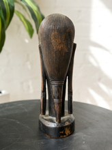 Load image into Gallery viewer, Image 8 of 13: A dark brown carved wooden sculpture of a stylized figure with a ribbed oval head and three legs, standing on a round dark tabletop against a light wall with soft shadows and a green plant leaf in the background.

