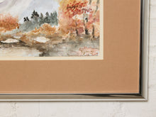 Load image into Gallery viewer, Image 7 of 8: A framed watercolor landscape print showing autumn trees and a stream, with a tan mat and a silver metal frame, mounted on a white brick wall.
