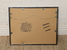 Load image into Gallery viewer, Image 8 of 8: The back of a framed picture with a tan cardboard backing, a dark metal frame, mounting clips, and some black scribbles on the surface, set against a white brick wall and woven rug.
