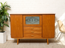 Load image into Gallery viewer, Image 1 of 27: Vintage mid-century modern teak high sideboard featuring a warm honey-brown wood finish, two large cabinet doors, three central drawers with curved metal handles, a decorative geometric glass-front compartment, and slender tapered wooden legs.
