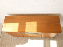 Load image into Gallery viewer, Image 4 of 27: A mid-century modern wooden console cabinet with a warm honey-amber finish, featuring a glass-front lower cabinet door, photographed from a high angle against a white brick wall and textured rug.
