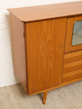 Load image into Gallery viewer, Image 8 of 27: A mid-century modern wooden sideboard cabinet with a warm honey-toned wood grain finish, featuring a cabinet door, drawers with chevron grain, and a small mirrored window, shown against a white brick wall and textured rug.
