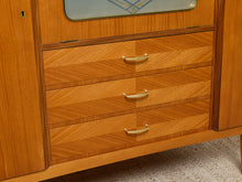 Load image into Gallery viewer, Image 13 of 27: Close-up of a mid-century modern wooden cabinet with three drawers featuring chevron-patterned wood grain and curved brass handles.
