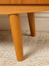 Load image into Gallery viewer, Image 17 of 27: Close-up of a light natural wood table leg and part of the tabletop, standing on a textured cream woven rug against a light wall.

