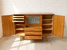 Load image into Gallery viewer, Image 21 of 27: A mid-century modern wooden credenza or sideboard cabinet with open doors, multiple drawers, open shelves, and a decorative glass panel in the center, shown against a white brick wall and light rug.
