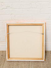 Load image into Gallery viewer, Image 8 of 8: The back of a square framed canvas or print, showing a light wood frame, white painted edges, and a hanging wire with safety clips, leaning against a white brick wall on a woven rug.
