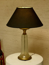 Load image into Gallery viewer, Image 4 of 18: A vintage table lamp with a black fabric conical shade, a clear glass flared base, and brass metal accents, sitting on a white cylindrical pedestal against a brick wall.
