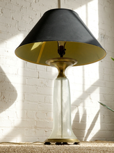 Load image into Gallery viewer, Image 5 of 18: A table lamp with a black fabric shade, a brass-toned neck and base, and a clear glass flared stem, set against a white brick wall with strong sunlight and shadows.
