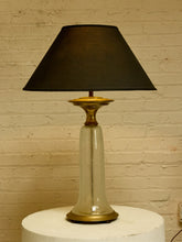 Load image into Gallery viewer, Image 8 of 18: A vintage-style table lamp with a black fabric conical shade, a clear glass flared base, and brass-toned metal accents, displayed on a white cylindrical pedestal against a beige brick wall.
