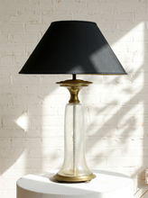 Load image into Gallery viewer, Image 7 of 18: A table lamp with a black fabric conical shade, a clear glass cylindrical base, and a brass-toned metal collar and base, displayed on a white pedestal against a white brick wall with dappled sunlight.
