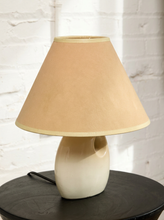 Load image into Gallery viewer, Image 3 of 14: A table lamp with a tapered beige fabric shade and a glossy cream-colored ceramic base, shown on a dark round table against a white brick wall background.
