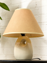 Load image into Gallery viewer, Image 5 of 14: A table lamp with a tapered beige fabric shade and a glossy ceramic base featuring a cream-to-beige gradient and a central cutout, set against a white brick wall.

