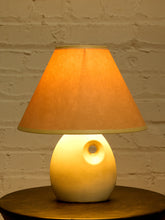 Load image into Gallery viewer, Image 8 of 14: A table lamp with a warm orange fabric shade and a glowing cream-colored ceramic base, set on a dark wooden surface against a white brick wall.
