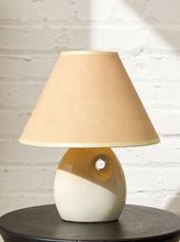 Load image into Gallery viewer, Image 7 of 14: A table lamp with a tapered beige fabric shade and a glossy ceramic base split horizontally into beige and off-white, sitting on a dark round table against a white brick wall.
