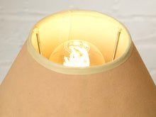 Load image into Gallery viewer, Image 9 of 14: Close-up of a warm beige fabric lampshade with a glowing light bulb visible inside, set against a light neutral background.
