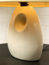 Load image into Gallery viewer, Image 13 of 14: A close-up of a ceramic table lamp base with a glossy white finish and a warm yellow-gold gradient on the upper half, shown on a dark tabletop against a light background.
