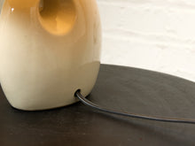 Load image into Gallery viewer, Image 14 of 14: Close-up of a cream-colored ceramic table lamp with a visible power cord exiting the base, resting on a dark tabletop against a white brick wall.
