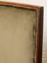 Load image into Gallery viewer, Image 8 of 15: Close-up of an upholstered chair corner showing a muted sage green velvet fabric cushion, a warm brown wooden frame, and a dark wood side rail against a light off-white background.
