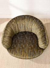 Load image into Gallery viewer, Image 6 of 16: A modern curved accent chair upholstered in a textured olive green fabric with vertical metallic gold and silver linear patterns, shown from a top-down angle on a woven rug against a white brick wall.
